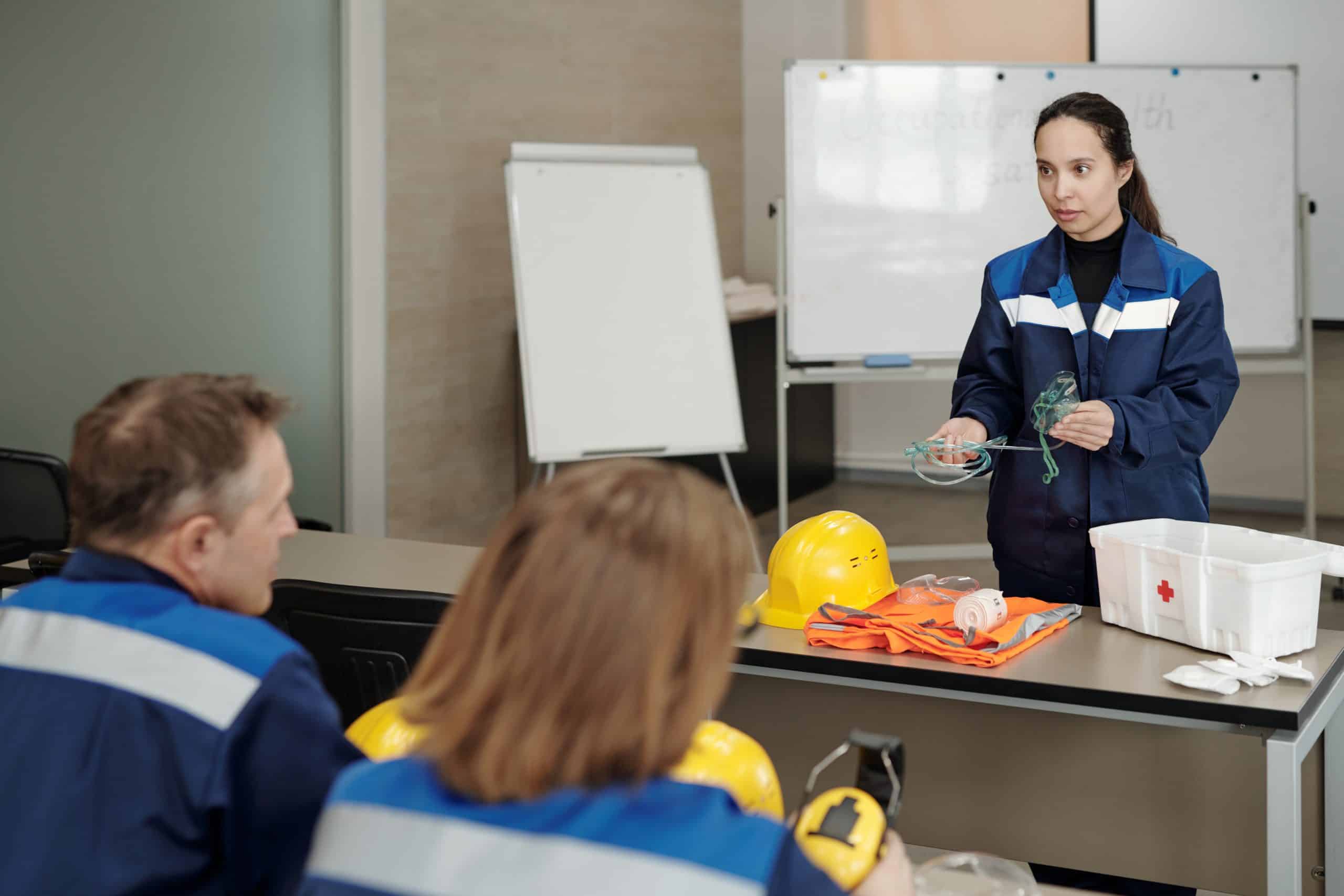 Female occupational safety instructor in blue jacket standing at table and holding oxygen mask while explaining how to give first aid