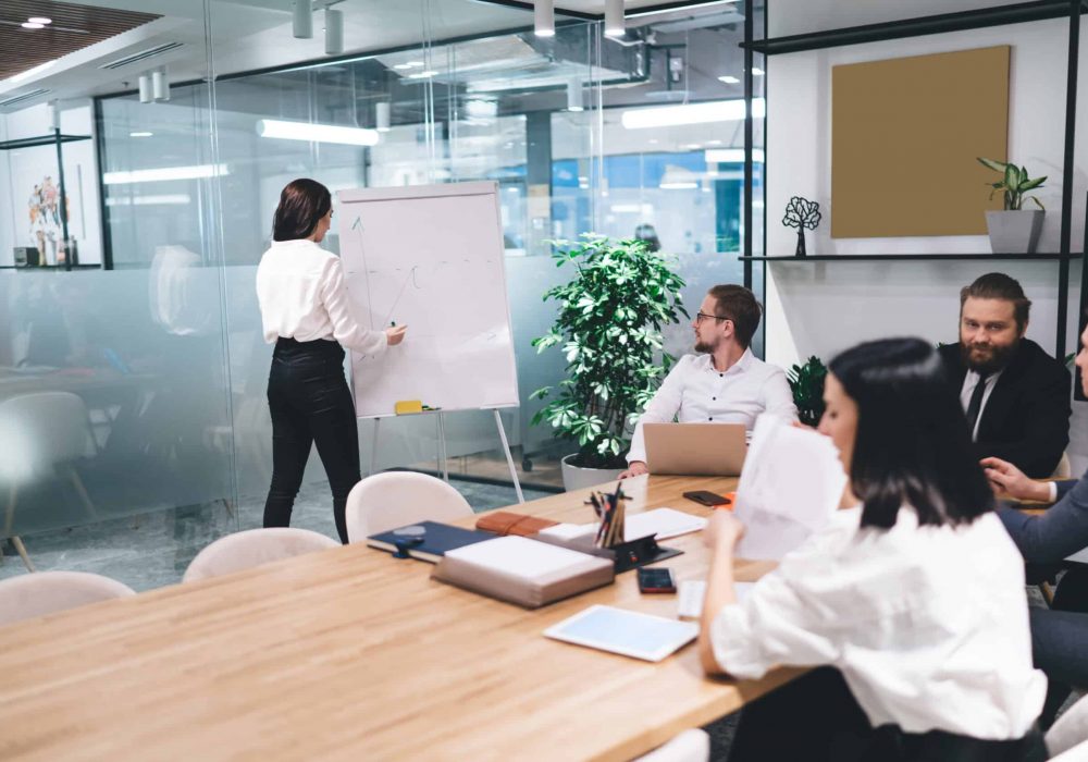 Busy female entrepreneur in formal clothes standing near whiteboard and taking notes with felt pen while working in modern meeting room and sharing business strategy with colleagues