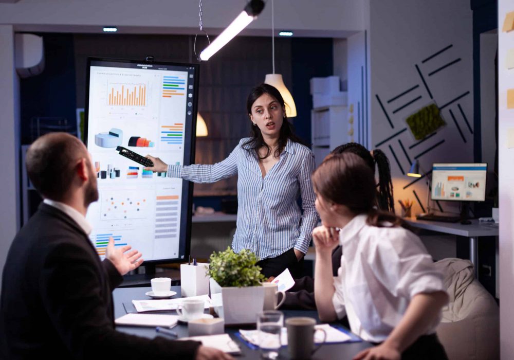 Workaholic focused businesswoman explaining management solution pointing strategy on monitor overworking in company business office meeting room. Multi-ethnic coworkers discussing ideas in evening